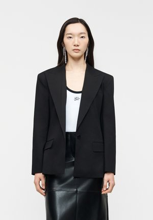 Ženska, oblečena v črni ohlapni blazer, belo rebrasto majico z majhnim logotipom, črno usnjeno krilo in dolge viseče uhane, stoji ob beli steni.