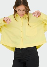 Gelbes Hemd mit Knöpfen und Tasche, entworfen in einem lockeren Schnitt mit breiten Ärmeln. Glatte Stoffqualität mit leichtem Glanz. Schwarze Hose.