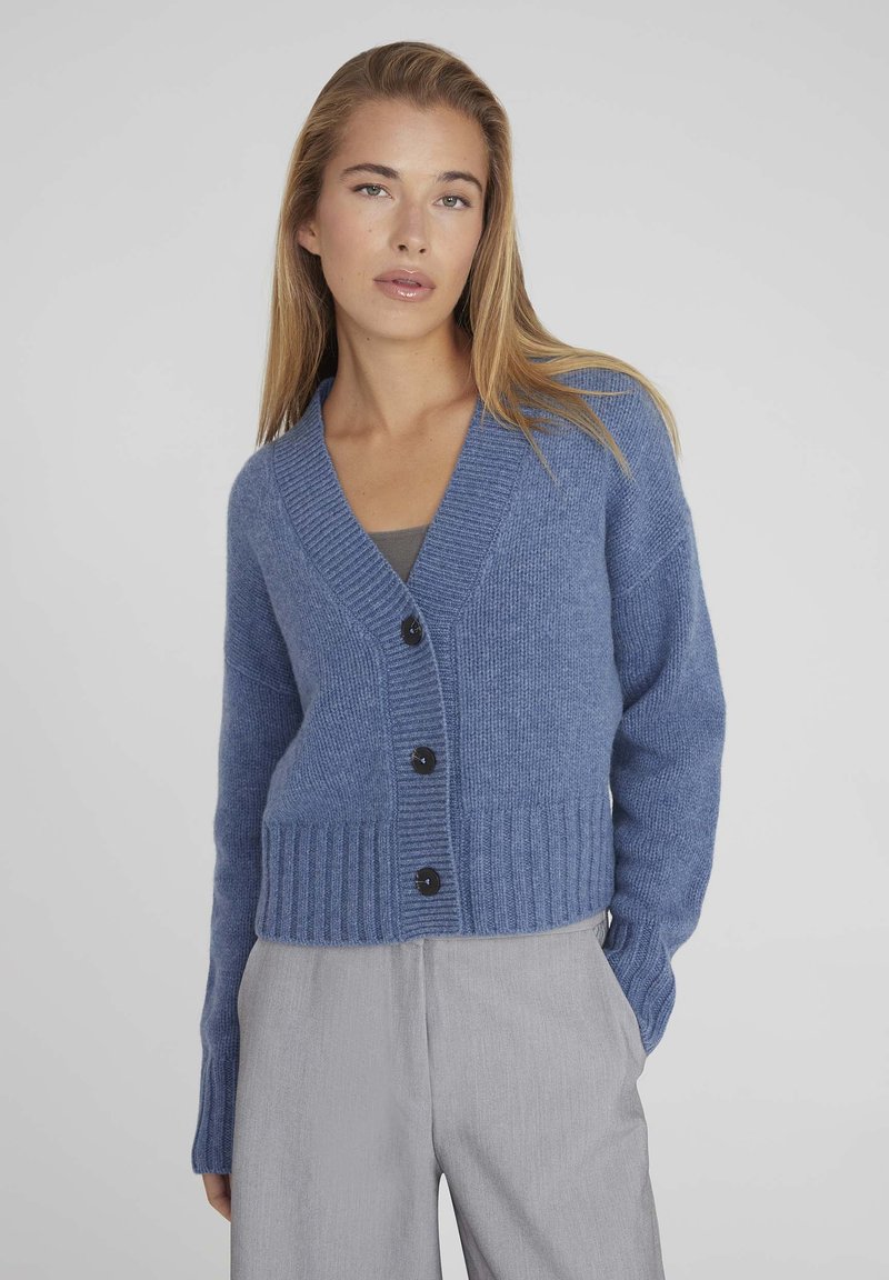 Cardigan en tricot bleu avec col en V, ourlet côtelé et six boutons noirs. Texture douce et coupe décontractée, associé à un pantalon gris clair.