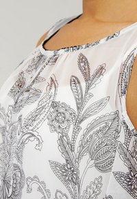 Haal white blouse, jossa on mustia kukka- ja lehtikuvioita, pyöreä kaula-aukko ja hihaton muotoilu. Teksturoitu kangas, jossa on herkkiä yksityiskohtia.