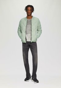 Lichtgroene gewatteerde bomberjack, grijze gestructureerde trui, donkere jeans en zwarte sneakers. Model staat met de handen in de zakken tegen een effen achtergrond.