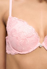 Κοντινό πλάνο ενός ατόμου που φοράει ροζ δαντελένιο σουτιέν με κεντήματα λουλουδιών και ένα μικρό μεταλλικό διακοσμητικό στο κέντρο μπροστά.