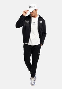 Zwarte zip-up jas met logo, witte t-shirt, zwarte cargobroek en kleurrijke sneakers met patroon, afgewerkt met een witte pet met tekst.
