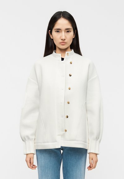 Veste blanche surdimensionnée avec un col montant, ornée de boutons ronds en bois et de poignets côtelés, portée sur un haut noir et un jean bleu.