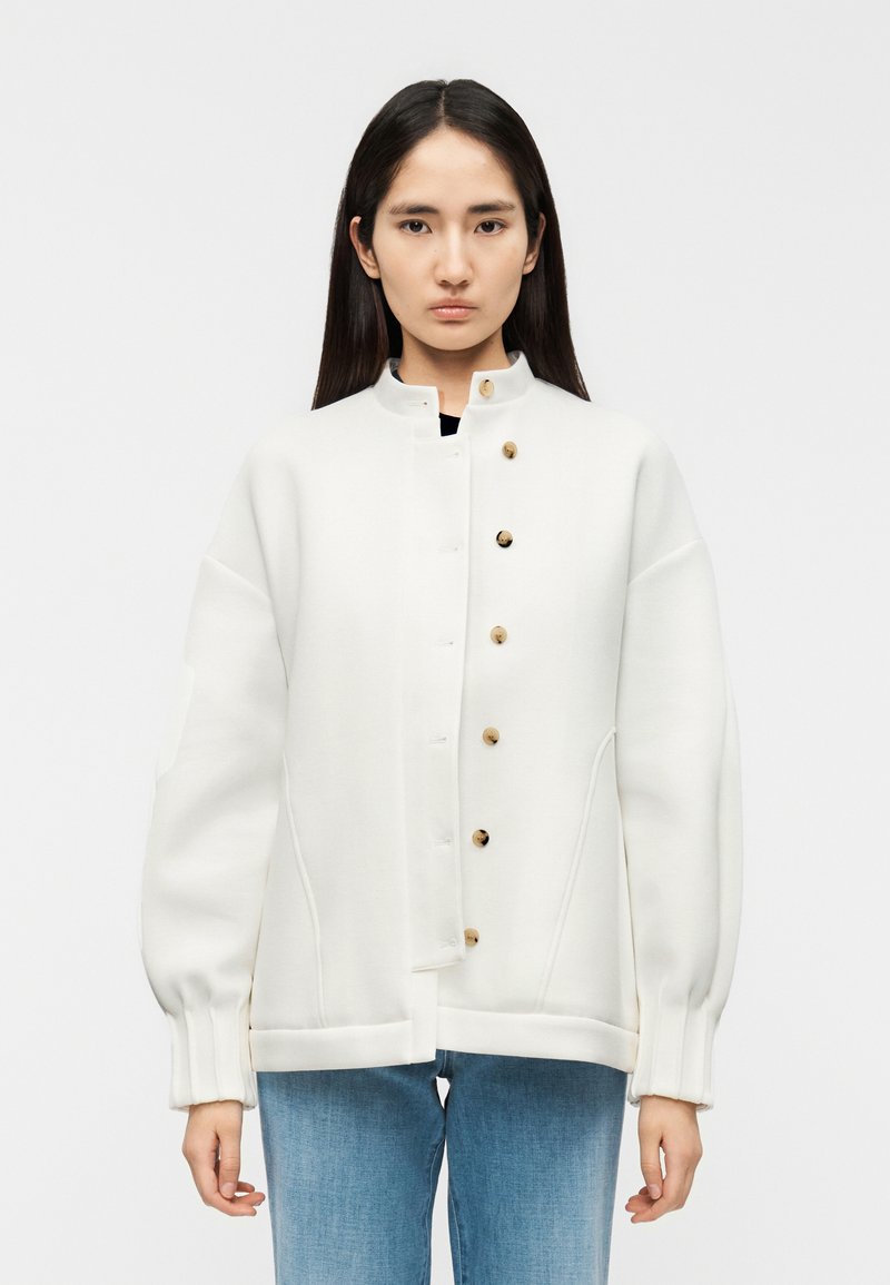 Λευκό, υπερμεγέθες μπουφάν με ψηλό γιακά, διακοσμημένο με στρογγυλά ξύλινα κουμπιά και ριμπ μανσέτες, φορεμένο πάνω από μαύρο τοπ και μπλε τζιν.