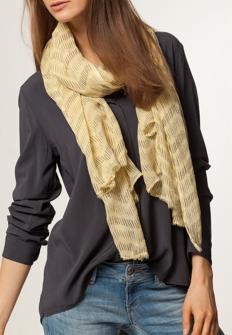 Femme portant une écharpe jaune clair à motifs sur un chemisier gris foncé à manches longues et un jean bleu, les cheveux lâchés et une posture détendue.