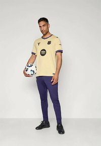 Beige voetbalshirt met paarse accenten en graphics, gecombineerd met paarse sportbroeken en zwarte voetbalschoenen, terwijl er een geschilderde voetbal wordt vastgehouden.