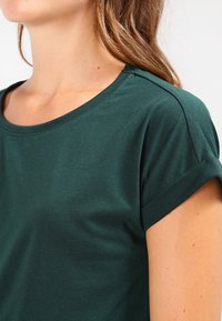 Tmavě zelené bavlněné tričko s kulatým výstřihem, krátkými ohrnutými rukávy a hladkým povrchem. Zaměření na detail ramene viditelný na obrázku.