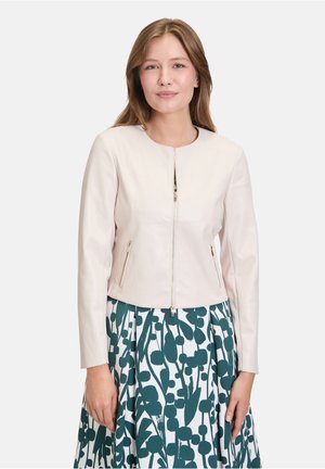Woman with long brown hair wearing a light beige zip-up jacket and a white skirt with large dark green abstract leaf patterns.
