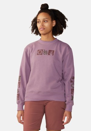 Femme aux cheveux naturellement bouclés portant un sweat violet avec des imprimés marron sur le thème de la nature et un short couleur rouille, debout sur un fond blanc.