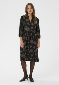 Rochie neagră cu un model floral bej, până la genunchi, decolteu în V, mâneci trei sferturi, talie strânsă, asortată cu pantofi slingback negri.