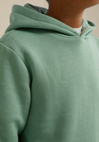 Felpa in cotone verde chiaro con una texture morbida, dotata di una tasca frontale e cappuccio con coulisse, mostrata dalla spalla in su.