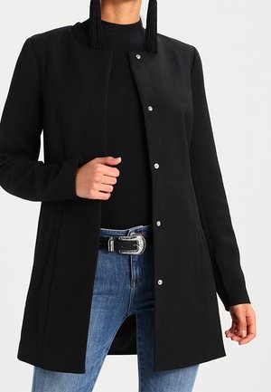 Femme portant un manteau noir à manches longues boutonné, un haut noir, un jean bleu avec une ceinture noire et une boucle argentée, ainsi que de longues boucles d'oreilles noires à pampilles.