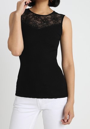 Kobieta ubrana w czarny, bezrękawnikowy top z prążkowanej dzianiny z koronkowym dekoltem, w białych spodniach, pozująca na jednolitym tle.
