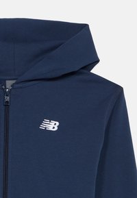 Tumši zilais džemperis ar rāvējslēdzēju, izgatavots no mīkstas auduma, ar priekšējo rāvējslēdzēju un baltu iešūtu logotipu kreisajā krūtī.