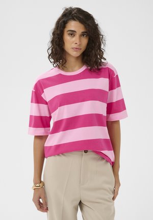 Vrouw met krullend haar, gekleed in een roze en lichtroze gestreept shirt met korte mouwen en beige broek, staand tegen een effen achtergrond.