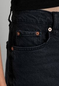 Los jeans de denim oscuro presentan una vista en primer plano de un bolsillo lateral con remaches de cobre, tela texturizada y una cinturilla suave, enfatizando el tono oscuro.