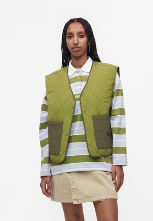 Kvinde med lange fletninger iført en grøn quiltet vest over en hvid og grønstribet langærmet poloshirt og beige nederdel.