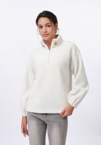 White fleece pullover with zippered collar, front buttons, and balloon sleeves; soft texture and relaxed fit; paired with light gray jeans.