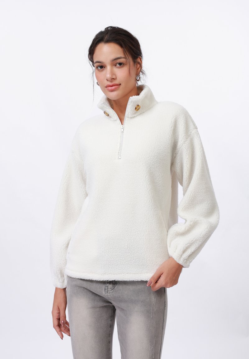 Pull en polaire blanc avec col zippé, boutons sur le devant et manches ballon ; texture douce et coupe décontractée ; associé à un jean gris clair.