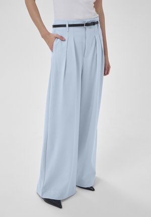 Osoba stojaca s jednou rukou v vrecku, oblečená v svetlomodrých širokých nohaviciach s vysokým pásom a záhybmi, čiernym opaskom, bielom topu a čiernych špicatých topánkach.