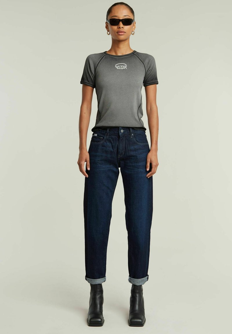 T-shirt grigia aderente con maniche corte e un logo, abbinata a jeans dritti blu scuro e stivaletti neri alla caviglia. Design semplice e casual.