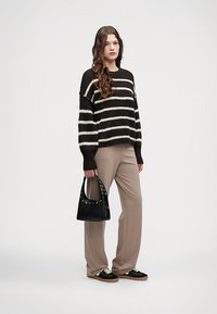 Randig svart och vit stickad tröja, beige byxor, svart väska med nitar och svarta sneakers med ljusa sulor. Avslappnad outfit på en modell.