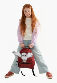 Zaino rosso con dettagli neri caratterizzato da un gufo cartone animato con le ali. La ragazza indossa una giacca zip lavanda, una maglietta bianca e jeans azzurri chiari.