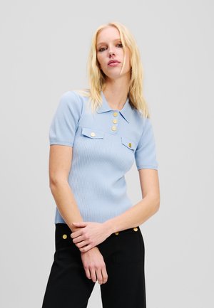 Polo en maille côtelée bleu clair à manches courtes, avec un col et trois boutons dorés à l'avant, associé à un pantalon noir taille haute.