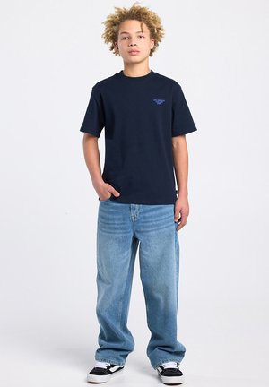 Giovane con capelli ricci in piedi che indossa una maglietta blu navy, jeans larghi azzurri e scarpe da ginnastica bianche e nere su uno sfondo semplice.