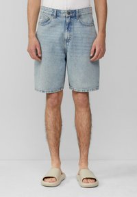 Shorts en denim bleu avec une coupe droite, cinq poches et un ourlet cousu. Portés avec des sandales à brides beige.
