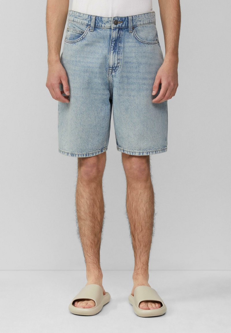 Shorts en denim bleu avec une coupe droite, cinq poches et un ourlet cousu. Portés avec des sandales à brides beige.