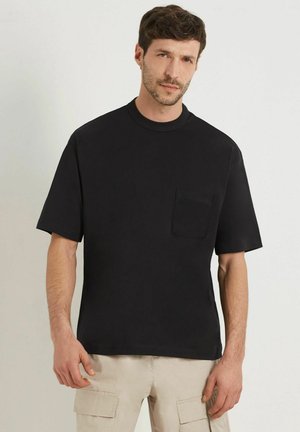 Tricou scurt din bumbac, de culoare neagră, cu mânecă scurtă, guler rotund, un singur buzunar pe piept și croială lejeră. Textură netedă, design minimalist.