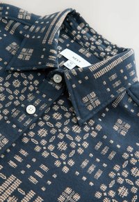 Tumesinine polosärk kreemilise geomeetrilise mustriga, millel on nööpidega kaelus ja kaks nööpi kaela juures. Silt ütleb "NEXT", suurus M.