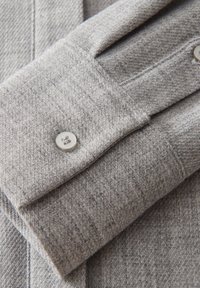Gros plan sur un poignet de chemise en laine grise avec un seul bouton blanc et des détails de couture fine visibles.