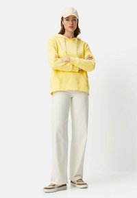 Geel hoodie met trekkoorden, crèmekleurige wijde broek en beige sneakers. Model staat met gekruiste armen en draagt een lichtgekleurde pet.