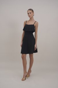 Robe noire sans manches, avec une superposition de volants au niveau du buste, taille cintrée, et jupe évasée. Associée à des talons dorés à lanières.