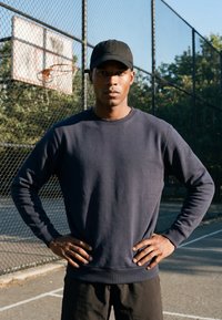 Jeune homme portant une casquette noire et un sweat-shirt foncé, debout les mains sur les hanches sur un terrain de basket en plein air près d'un panier et d'une clôture en chaîne.