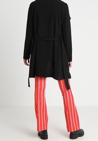 Černý dlouhý kardigan s texturovanou tkaninou a zavazovacím páskem, v kombinaci s červenými pruhovanými rozšířenými kalhotami. Viditelné černé boty s robustní podrážkou.