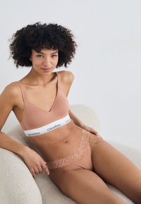 Bralette en coton marron clair avec une large bande Calvin Klein et un string en dentelle assorti sur un modèle assis devant un fond neutre.