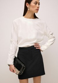 Camisa blanca de manga larga con detalles de botones, combinada con una falda negra. Clutch negro de piel de serpiente sostenido en la mano. Texturas de tela suaves.