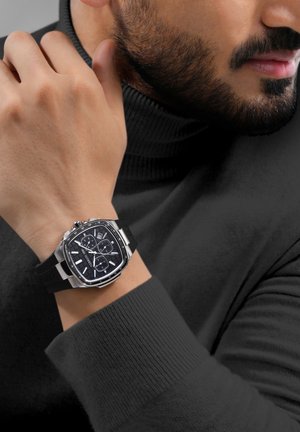 Montre chronographe noire avec un boîtier carré en acier inoxydable, un bracelet en caoutchouc noir, des index horaires blancs et un cadran sombre texturé. Portée sur un poignet.