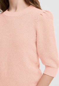 Ružový pletený sveter s textúrovaným povrchom, okrúhlym výstrihom a balónovými rukávmi po 3/4 dĺžke. Materiál sa zdá byť mäkký a ľahký.