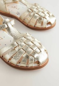Gyldne metalliske vævede sandaler med åben tå, flad sål og polstret indersål med blomsterprint. Glat tekstur og justerbar rem.