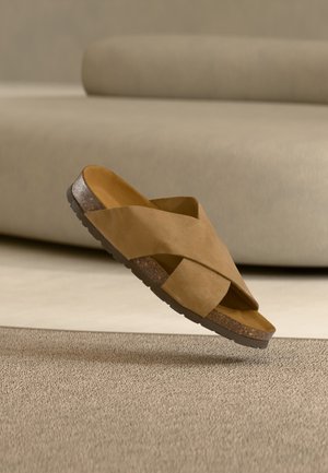 Brązowa zamszowa klapka ze skrzyżowanymi paskami i korkową wkładką, unosząca się nad teksturowanym beżowym dywanem w pomieszczeniu o neutralnej kolorystyce.