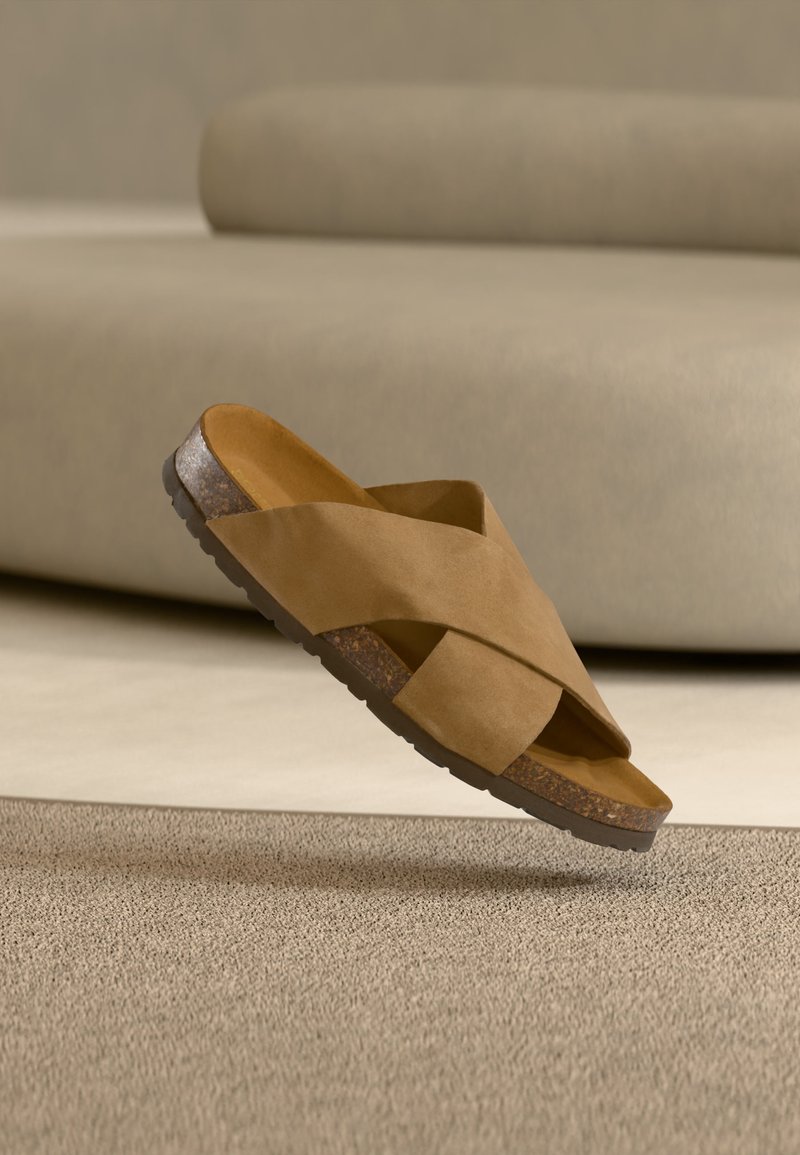 Καφέ σανδάλι slide από κ suede με σταυρωτούς ιμάντες και πάτο από φελλό, αιωρείται πάνω από μια υφασμάτινη μπεζ μοκέτα σε ένα δωμάτιο με ουδέτερους τόνους.