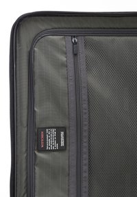 Interior de maleta verde oscura con un bolsillo de malla, compartimentos con cremallera y una etiqueta con instrucciones de cuidado. Textura suave y diseño estructurado.