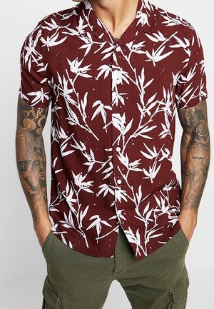 Chemise à manches courtes couleur bordeaux avec un motif de feuilles de bambou blanches, design à boutons, et en tissu léger. Assortie avec un pantalon cargo vert.