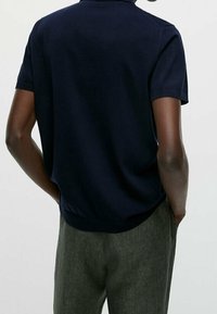 T-shirt polo bleu marine à manches courtes vu de dos ; il présente un col en ribbed, une texture lisse et une coupe décontractée. Associé à un pantalon gris.