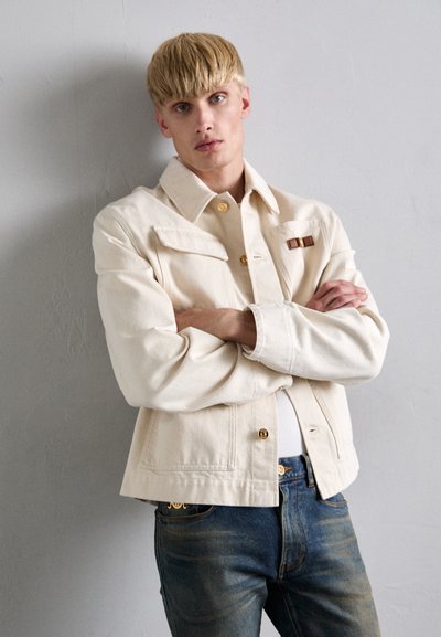 Beige denim takki, jossa on kauluksellinen muotoilu, etupainonappikiinnitys ja rintataskut. Käytetään sinisten farkkujen päällä, joissa on kultaiset yksityiskohdat.